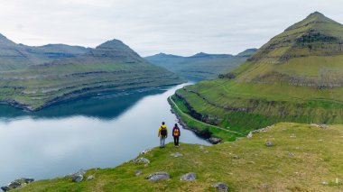 İki yürüyüşçü, aşağıdaki fiyordun sakin sularına tepeden bakan çarpıcı yeşil bir vadinin kenarında duruyor. Faroe Adaları 'nın engebeli zirveleri dramatik bir zemin oluşturuyor, Gonguturur Hvithamar. 