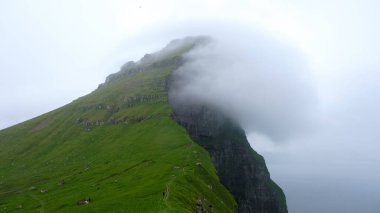 Faroe Adaları 'ndaki çarpıcı bir dağ zirvesi, kısmen dönen bulutlarla çevrili yemyeşil tepelerin üzerinde dramatik bir şekilde yükselir. Bu manzara, bu uzak hedefin vahşi güzelliğini yakalar..