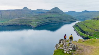 İki yürüyüşçü Faroe Adaları 'nın nefes kesici manzarasına bakan kayalık bir çıkıntıda duruyor. Sakin sular, uzaktaki sivri dağları yansıtır ve sakin bir atmosfer yaratır..