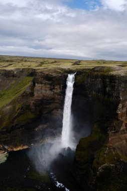 Görkemli Haifoss şelalesi sarp kayalıkların üzerinden zarif bir şekilde akar, yemyeşil manzaralarla çevrilidir. Dramatik gökyüzü bu sakin İzlanda doğal harikasına derinlik katıyor..