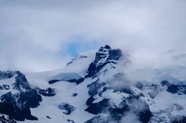 Svinafellsjokull Buzulu, yükselen kayalık tepeler tarafından çerçevelenmiş mavi ve beyaz tonlarda parıldıyor. Bulutlar sakin bir şekilde sürükleniyor İzlanda 'da nefes kesici vahşi doğada mistik bir atmosfer yaratıyor..