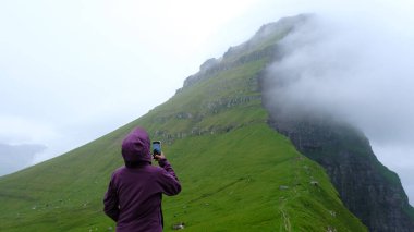 Mor ceketli bir gezgin yemyeşil yamaçlarda duruyor, Faroe Adaları 'nın dramatik uçurumlarını yakalıyor, zirvenin etrafında dönen sis, Kallur Deniz feneri, Kalsoy Adası