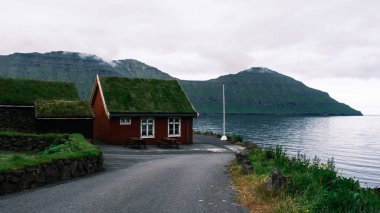 Faroe Adaları 'nın dingin sularının yanında, dramatik dağ manzaralarıyla çevrili pitoresk yeşil çatılı bir ev bulunuyor. Bulutlu gökyüzü sakin ortama kasvetli bir atmosfer ekler..