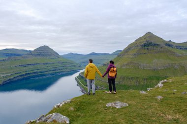 İki yürüyüşçü, Faroe Adaları 'ndaki Gonguturur Hvithamar Patikası boyunca uzanan büyüleyici manzara karşısında el ele tutuşuyor. Bereketli yeşil tepeler ve sakin sular sakin bir atmosfer yaratır..