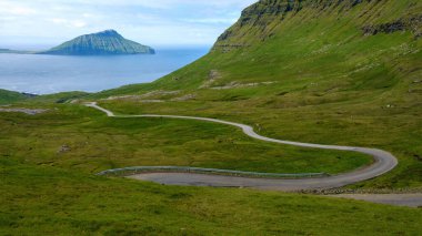 Faroe Adaları 'nın canlı yeşil tepelerinde kıvrılan dolambaçlı bir yolun nefes kesici manzarası. Arka planda dingin okyanus ışıldıyor, ufukta görkemli uçurumlar var..