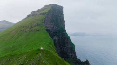 Faroe Adaları 'nda, denizin üzerindeki dik uçurum kulesinde, yemyeşil çimlere gömülmüş parlak bir deniz feneri, sisli bir atmosferle kaplanmış, Kallur Deniz feneri Kalsoy Adası.
