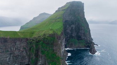 Faroe Adaları 'ndaki okyanustan sisle kaplı görkemli uçurumlar dramatik bir şekilde yükseliyor. Bu çarpıcı manzara doğanın özünü yansıtıyor ham güzellik, Kallur Deniz Feneri Kalsoy Adası