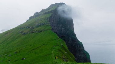 Tek başına bir yürüyüşçü, yüksek uçurumlarla çevrili, yeşil yamaçlardan geçer ve sisle örtülmüş, vahşi güzelliği ve engebeli araziyi yakalar, Kallur Deniz feneri Kalsoy Adası
