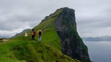 İki maceracı, Faroe Adaları 'nın çarpıcı kayalıklarına ve yemyeşil yeşilliğine hayran olmak için durdular. Dramatik arazi okyanusla buluşuyor, Kallur Deniz Feneri, Kalsoy Adası