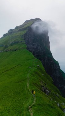 Bir çift, verimli yeşil manzaralar ve sürüklenen bulutlarla çevrili, Faroe Adaları 'nın sarp kayalıkları boyunca dolambaçlı bir yoldan geçer. Kallur Deniz Feneri Kalsoy Adası