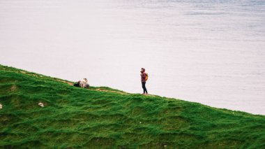 Bir gezgin otlak bir tepede duruyor, sakin suyun ve Faroe Adaları 'nın verimli tepelerinin nefes kesici manzarasının tadını çıkarıyor. Kallur Deniz Feneri, Kalsoy Adası