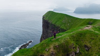 Canlı bir maceracı Faroe Adaları 'ndaki çarpıcı yeşil bir uçurumun kenarında muzaffer bir şekilde duruyor. Kallur Deniz Feneri Kalsoy Adası