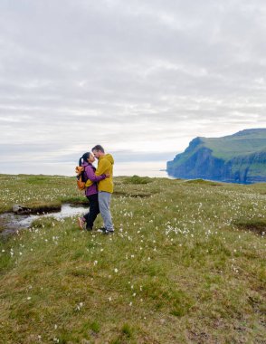 Bir çift, Faroe Adaları 'ndaki çimenli bir alanda etraflarında canlı kır çiçekleriyle hassas bir anı paylaşıyor. Yumuşak bulutlu bir gökyüzünün altında arka planda görkemli kayalıklar yükselir. Eidiskollur Eidi 