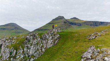 Gonguturur Hvithamar Patikası 'ndaki kayalıkların tepesinde iki yürüyüşçü duruyor. Etrafı yemyeşil tepeler ve Faroe Adaları' nın dramatik uçurumlarıyla çevrili..