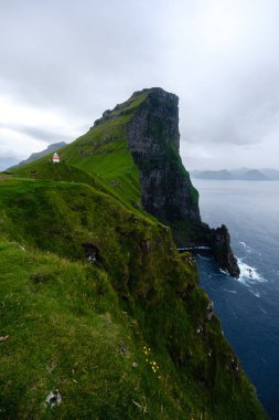 Faroe Adaları 'ndaki Atlantik Okyanusu' ndan yükselen sarp kayalıklar yemyeşil çimlerle kaplıdır. Bulutlu gökyüzü Kallur Deniz feneri Kalsoy Adası 'na mistik bir cazibe katıyor.