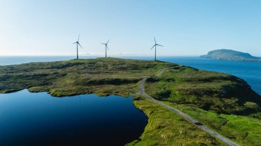 Rüzgâr türbinleri, sakin sular ve açık bir gökyüzüyle çevrili Faroe Adaları 'ndaki çimenli tepelerde dimdik durur. Bu pitoresk manzara doğanın uyumunu ve yenilenebilir enerjiyi gösteriyor..