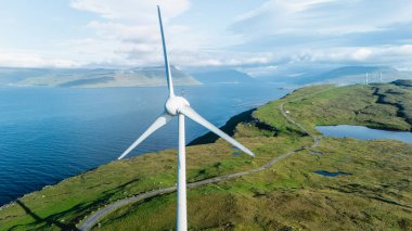 Berrak yeşil tepeler ve sakin sularla çevrili sakin Faroe Adaları 'nın arka planında heybetli bir rüzgar türbini dimdik ayakta duruyor..