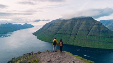 İki yürüyüşçü Faroe Adaları 'ndaki Klakkur noktasından gelen çarpıcı manzaraya hayran. Yumuşak tepeler sakin sularla buluşur ve bu uzak cennetin özünü yansıtan pitoresk bir manzara yaratır..