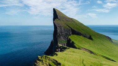Ziyaretçiler Faroe Adaları 'nda nefes kesici manzaraların keyfini çıkarırken Majestic Mylingur kayalıkları Atlantik' ten dramatik bir şekilde yükseliyor. Bereketli yeşil manzaralar açık bir günde derin mavi denizle tezat oluşturur..