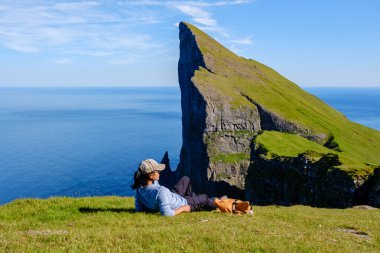 Çimenli bir çıkıntıya kurulmuş bir gezgin, Faroe Adaları 'ndaki dramatik Mylingur uçurum kenarının berrak gökyüzü ve berrak okyanus sularının ötesine uzanan nefes kesici manzarasının tadını çıkarıyor..