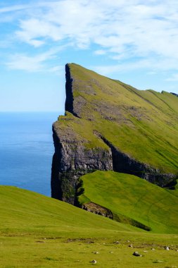 Faroe Adaları 'nın nefes kesici manzaraları yükselen uçurumları ve yemyeşil tepeleri gözler önüne seriyor. Bu büyüleyici manzara, saf güzelliği berrak mavi gökyüzünün altında sakin bir denizin arka planına karşı sergiliyor..