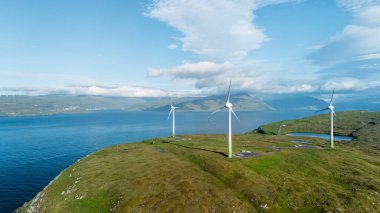 Görkemli rüzgar türbinleri, çimli bir tepenin üzerinde dikilip Faroe Adaları 'ndaki esintili enerjiyi yakalıyorlar. Aşağıdaki sakin sular, canlı mavi gökyüzünü yansıtıyor, resim gibi bir manzara yaratıyor..