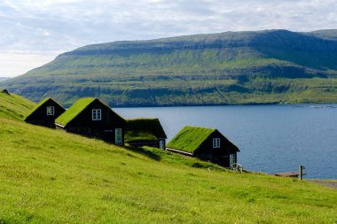 Geleneksel çim çatılı evler, güneşli bir günde Faroe Adaları 'nın eşsiz mimarisini ve nefes kesici manzarasını gözler önüne sererek, sakin suların yanındaki yemyeşil araziyi dolduruyor. Bour Köyü