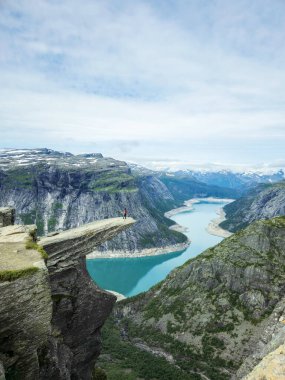 Cesur bir yürüyüşçünün dramatik bir uçurumun kenarında durduğu geniş bir manzara. Aşağıda, bir fiyordun kristal berrak suları çevresindeki görkemli dağları yansıtır. Norveç 'te Trolltunga yürüyüşü
