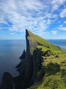 Faroe Adaları 'ndaki Mylingur' un nefes kesici uçurumlarını keşfedin. Dramatik manzaralar ve canlı yeşil çimlerle çevrili bir yürüyüşçü kenarda durur. Aşağıdaki okyanus ışıldıyor, güzelliği arttırıyor..