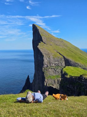 Faroe Adaları 'ndaki Mylingur yürüyüşünden nefes kesici manzaralar, yemyeşil çimlerde dinlenen bir yürüyüşçüyü sergiliyor, dramatik uçurumlar ve açık mavi bir gökyüzünün altında okyanus..