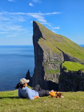 Faroe Adaları 'ndaki Mylingur' da bir yürüyüş sırasında sükuneti tecrübe edin. Bir yürüyüşçü yemyeşil kayalıklara yaslanır, engin okyanusa ve açık mavi gökyüzü altındaki dramatik kaya oluşumlarına bakar..