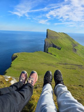İki yürüyüşçü çimenli bir çıkıntıda oturup dramatik Mylingur dağının ve Faroe Adaları 'nın etrafındaki okyanusun nefes kesici manzarasının tadını çıkarıyorlar. Mükemmel bir açık hava macera merkezi..