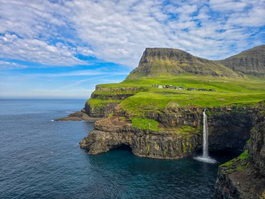 Faroe Adaları 'ndaki Gasadalur köyünün yemyeşil kayalıklarıyla çevrili Atlantik Okyanusu' na çarpıcı bir şekilde batarken Mulafossur Şelalesi 'nin nefes kesici güzelliğini deneyimleyin..