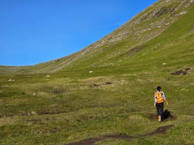 Bir yürüyüşçü, canlı mavi bir gökyüzü altında dramatik tepelerle çevrili Faroe Adaları 'ndaki Mylingur patikasının yemyeşil manzarasını dolaşır. Doğanın güzelliği maceracıları cezbeder..