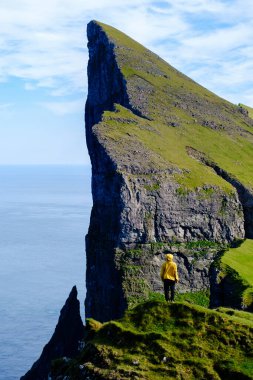 Bir gezgin, Faroe Adaları 'nın dramatik uçurumlarında yüksek kaya oluşumlarına ve engin okyanusa hayret ederek duruyor. Bereketli yeşil tepeler denizle buluşuyor, büyüleyici bir doğal manzara yaratıyor..