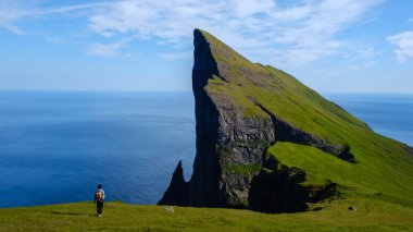 Yalnız bir yürüyüşçü yemyeşil bir yamaçta duruyor ve Faroe Adaları 'nın dramatik Mylingur uçurumlarına bakıyor. Dalgalı mavi deniz, yükselen kaya oluşumu ve berrak gökyüzü ile çok güzel tezat oluşturuyor..