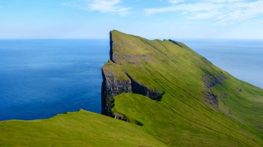 Mylingur uçurumları, Faroe Adaları 'ndaki yemyeşil çimlerle çevrili derin mavi okyanusun üzerinde dramatik bir şekilde yükselir. Açık gökyüzü nefes kesen kıyı manzarasını güçlendirir, keşifleri davet eder..