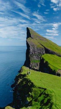 Majestic Mylingur kayalıkları Faroe Adaları 'ndaki pırıl pırıl okyanusun üzerinde çarpıcı bir şekilde yükseliyor. Yürüyüşçüler, engin mavi gökyüzü ve tüylü bulutlara karşı çarpıcı yeşil araziyi keşfediyorlar.
