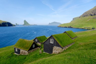 Faroe Adaları 'nın yemyeşil yeşilliklerine yerleşen geleneksel çim çatılı kulübeler, mavi suların ve uzak adaların zeminine karşı manzarayı noktalıyor. Bour Köyü
