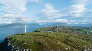 Faroe Adaları 'nın engebeli kıyı şeridinin çarpıcı hava manzarası çimenli bir tepede rüzgar türbinleri sergiliyor..