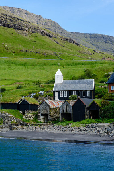 Nestled by the shoreline, charming black and white church stand against the vivid green hills of the Faroe Islands. Crisp blue waters gently lap the beach, Bour Village