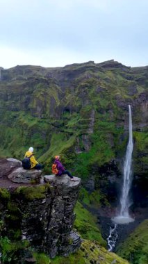 Ziyaretçiler, Mulagljufur Kanyonu 'ndaki kayalık bir uçurumun tepesinde, yemyeşil tepeler ve çağlayan şelalelerle çevrili olarak oturuyorlar. Doğal güzellik, sakin bir manzara ve maceraperest bir keşifle büyülenir..