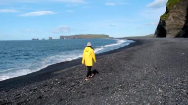 Asyalı bir kadın, İzlanda 'daki Reynisfjara Sahili' nin eşsiz siyah kumları boyunca yürüyor. Bu kıyı taşı çarpıcı kaya oluşumları ve büyüleyici okyanus dalgaları, davetkar keşif ve sükunet içerir..