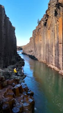 İzlanda 'daki Studlagil Kanyonu' nun nefes kesici güzelliğini keşfedin. Yüksek bazalt sütunların sakin sularla buluştuğu yer. Bir çift erkek ve kadın nefes kesen bir ortamda sanatın doğasına tanıklık ediyorlar..