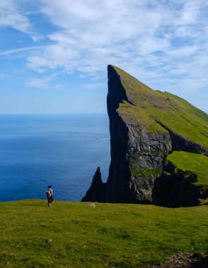 Faroe Adaları 'ndaki yemyeşil Mylingur uçurumları boyunca yürüyen yalnız bir yürüyüşçü, denizden keskin bir şekilde yükselen dramatik bir kaya oluşumundan büyülenmiştir. Parlak manzara açık gökyüzü tarafından aydınlatılır..