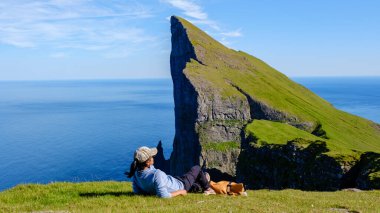 Bir gezgin, canlı yeşil tepeler ve uçsuz bucaksız okyanuslarla Faroe Adaları 'ndaki bir uçurumun nefes kesici manzarasının tadını huzur içinde çıkarır. Güneşli gökyüzünün altında meraklı bir köpek uzanır.