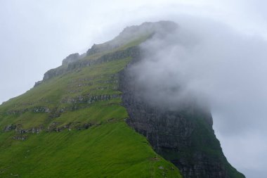 Yükselen yeşil uçurumlar yemyeşil bitki örtüsüyle kaplı, sisle kaplı gökyüzüne doğru uzanıyor. Kallur Deniz Feneri, Kalsoy Adası