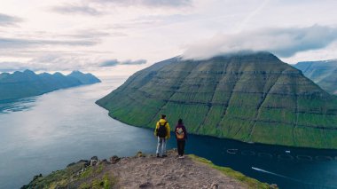 İki maceracı Klakkur 'un engebeli bir uçurumunun kenarında durup Faroe Adaları' nın nefes kesici manzarasının tadını çıkarıyorlar. Bereketli yeşil tepeler aşağıdaki dramatik kıyı şeridi ve sakin sularla tezat oluşturuyor..