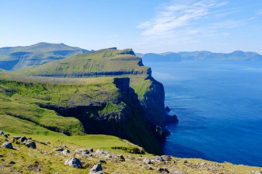 Görkemli uçurumlar, Faroe Adaları 'nın kıyı şeridi boyunca açık mavi bir gökyüzünün altında uzanır. Canlı yeşil tepeler ışıl ışıl Atlantik 'le buluşarak nefes kesici bir doğal manzara yaratıyor..