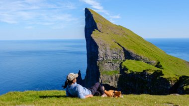 Bir gezgin, canlı yeşil tepeler ve uçsuz bucaksız okyanuslarla Faroe Adaları 'ndaki bir uçurumun nefes kesici manzarasının tadını huzur içinde çıkarır. Güneşli gökyüzünün altında meraklı bir köpek uzanır.